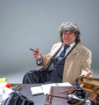 The senior man in cloak as detective or mafia boss writing in a notebook with pen at table. Studio shot on gray in retro stile