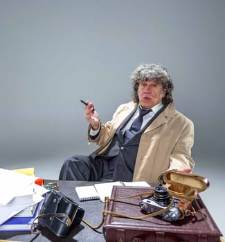 The senior man in cloak as detective or mafia boss writing in a notebook with pen at table. Studio shot on gray in retro stile