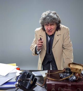 The senior man in cloak as detective or mafia boss writing in a notebook with pen at table. Studio shot on gray in retro stile
