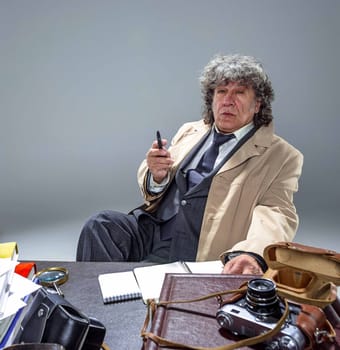 The senior man in cloak as detective or mafia boss writing in a notebook with pen at table. Studio shot on gray in retro stile