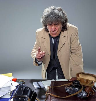 The senior man in cloak as detective or mafia boss writing in a notebook with pen at table. Studio shot on gray in retro stile