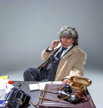 The senior man in cloak as detective or mafia boss writing in a notebook with pen at table. Studio shot on gray in retro stile