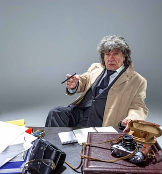 The senior man in cloak as detective or mafia boss writing in a notebook with pen at table. Studio shot on gray in retro stile