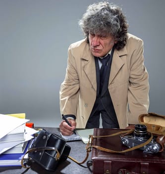 The senior man in cloak as detective or mafia boss writing in a notebook with pen at table. Studio shot on gray in retro stile
