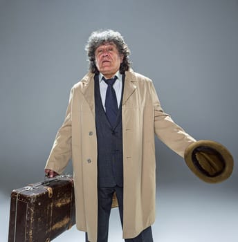 The senior man in cloak as detective or mafia boss. Studio shot on gray in retro stile. Mature man with hat and suitcase