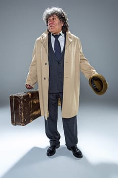 The senior man in cloak as detective or mafia boss. Studio shot on gray in retro stile. Mature man with hat and suitcase