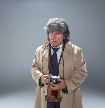 The senior man in cloak as detective or mafia boss with photo camera. Studio shot on gray in retro stile