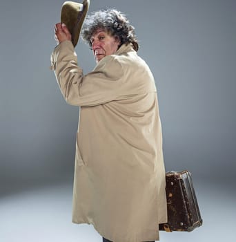 The senior man in cloak as detective or mafia boss. Studio shot on gray in retro stile. Mature man with hat and suitcase