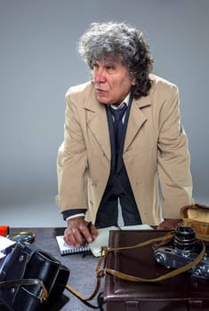 The senior man in cloak as detective or mafia boss writing in a notebook with pen at table. Studio shot on gray in retro stile