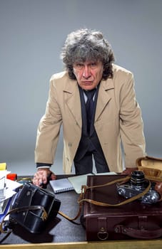 The senior man in cloak as detective or mafia boss writing in a notebook with pen at table. Studio shot on gray in retro stile