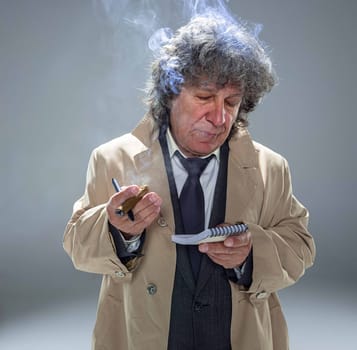 The senior man in cloak as detective or mafia boss writing in a notebook with pen. Studio shot on gray in retro stile