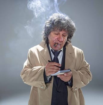 The senior man in cloak as detective or mafia boss writing in a notebook with pen. Studio shot on gray in retro stile