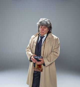 The senior man in cloak as detective or mafia boss with photo camera. Studio shot on gray in retro stile