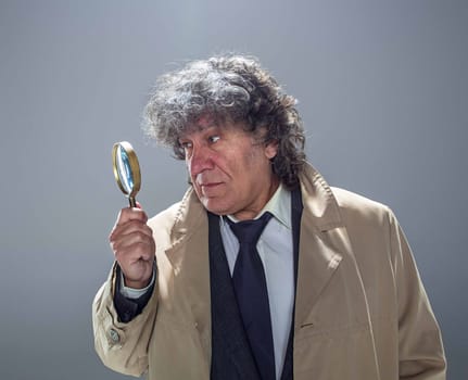 The senior man in cloak as detective or mafia boss with With a magnifying glass. Studio shot on gray in retro stile. Concept of Search