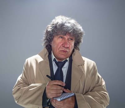 The senior man in cloak as detective or mafia boss writing in a notebook with pen. Studio shot on gray in retro stile