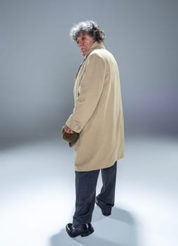 The back view of senior man in cloak as detective or mafia boss. Studio shot on gray in retro stile. Mature man with hat and suitcase