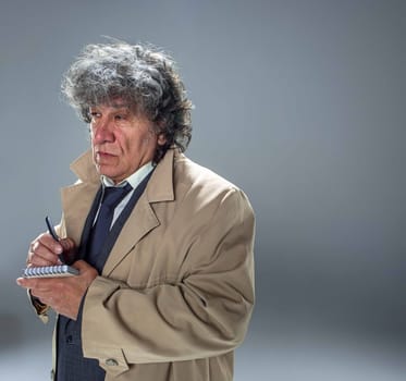 The senior man in cloak as detective or mafia boss writing in a notebook with pen. Studio shot on gray in retro stile