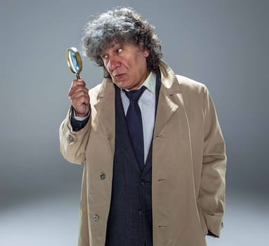 The senior man in cloak as detective or mafia boss with With a magnifying glass. Studio shot on gray in retro stile. Concept of Search