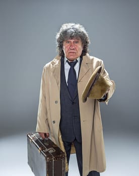 The senior man in cloak as detective or mafia boss. Studio shot on gray in retro stile. Mature man with hat and suitcase