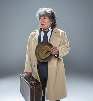 The senior man in cloak as detective or mafia boss. Studio shot on gray in retro stile. Mature man with hat and suitcase