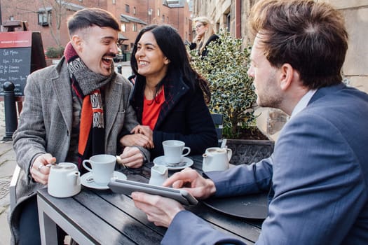 Happy couple are sitting with a financial advisor at a cafe. He is showing them a digital tablet with information and they are laughing while drinking tea.