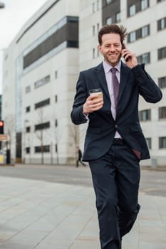 Businessman is walking to work in the city. He is holding a coffee cup and is talking on the phone.