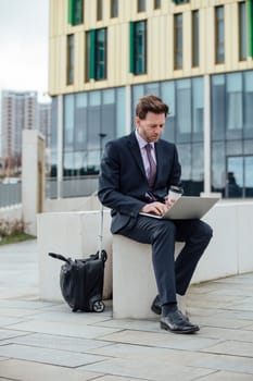 Businessman is working in the city. He is using a laptop and drinking a coffee.