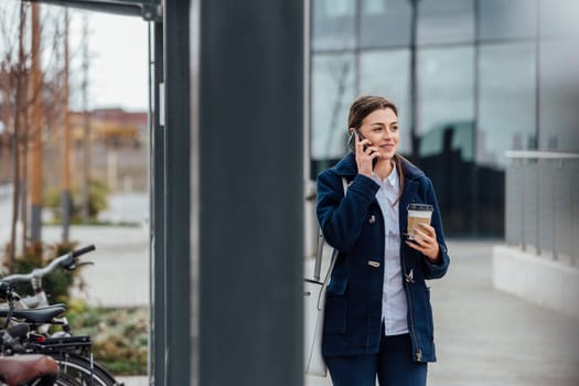 Businesswoman is walking to work in the city. She is holding a coffee cup and is talking on the phone.