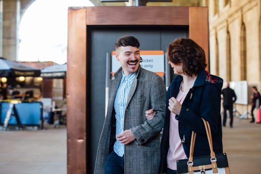 Couple are walking into the train station with their luggage. Theya re smiling and have their arms linked as they walk.