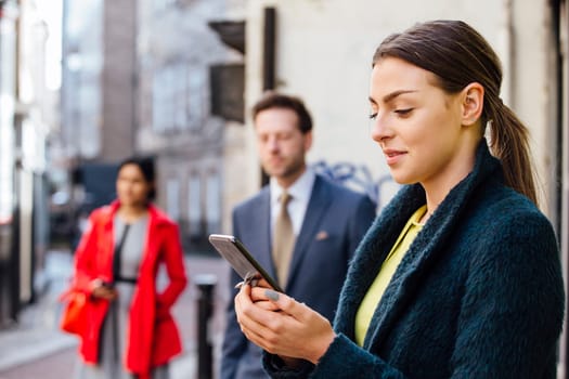 Young businesswoman is using her smartphone to book a taxi to her workplace. There is another man waiting at the taxi rank who can be seen defocussed in the background, as well as another woman who is walking up the street.