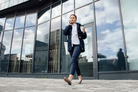 Young businesswoman is leaving her office building with a cup of tea. She is talking on the phone to someone as she walks.