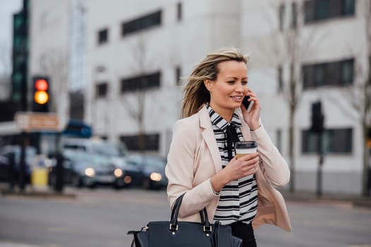 Businesswoman is walking to work in the city. She is holding a coffee cup and is talking on the phone.