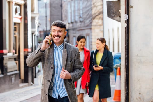 Businessman is walking through the city to go to work. He is well dressed and is talking on the phone, while two women can be seen in the background walking together while lookin at a smart phone.