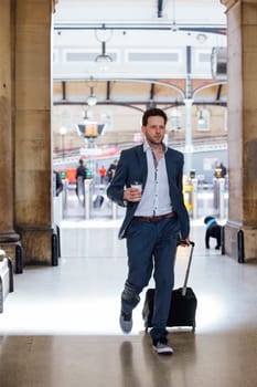 Businessman is in a hurry as he leaves the train station to commute the work. He is carrying a coffee and pulling a suitcase.