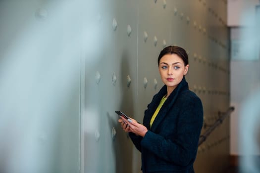 Businesswoman is standing in her workplace, looking out of her office window with a smart phone in her hands.
