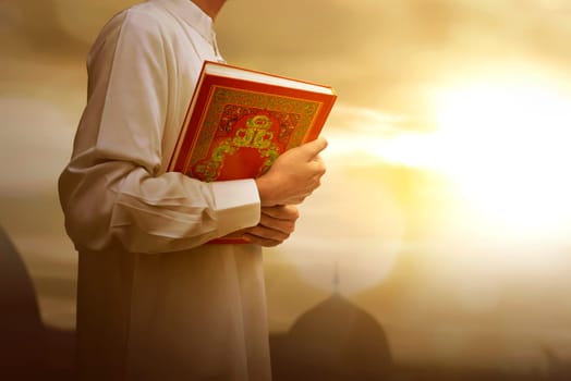Muslim man in traditional dress holding holy book Koran during sunset background