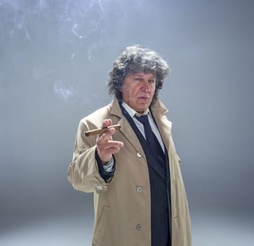 The senior man in cloak with cigar as detective or mafia boss. Studio shot on gray in retro stile