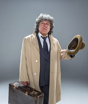 The senior man in cloak as detective or mafia boss. Studio shot on gray in retro stile. Mature man with hat and suitcase