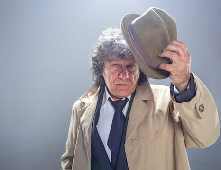 The senior man in cloak as detective or mafia boss. Studio shot on gray in retro stile. Mature man with hat and suitcase