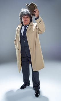 The senior man in cloak as detective or mafia boss. Studio shot on gray in retro stile. Mature man with hat and suitcase