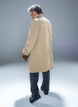 The back view of senior man in cloak as detective or mafia boss. Studio shot on gray in retro stile. Mature man with hat and suitcase
