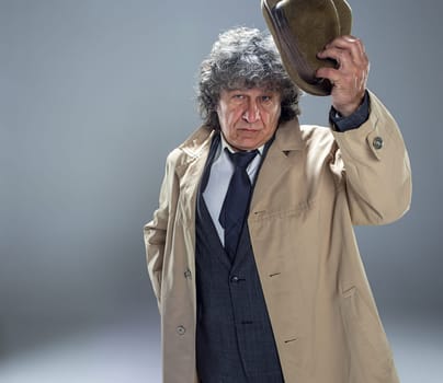 The senior man in cloak as detective or mafia boss. Studio shot on gray in retro stile. Mature man with hat and suitcase