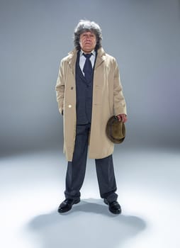 The senior man in cloak as detective or mafia boss. Studio shot on gray in retro stile. Mature man with hat and suitcase