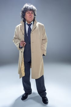 The senior man in cloak with cigar as detective or mafia boss. Studio shot on gray in retro stile