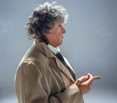 The senior man in cloak with cigar as detective or mafia boss. Studio shot on gray in retro stile