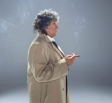 The senior man in cloak with cigar as detective or mafia boss. Studio shot on gray in retro stile
