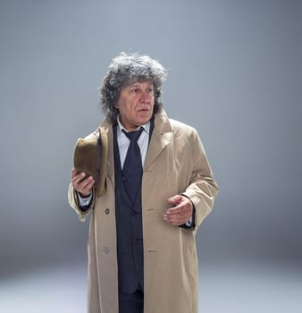 The senior man in cloak as detective or mafia boss. Studio shot on gray in retro stile. Mature man with hat and suitcase