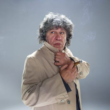The senior man in cloak with cigar as detective or mafia boss. Studio shot on gray in retro stile