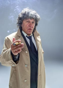 The senior man in cloak with cigar as detective or mafia boss. Studio shot on gray in retro stile
