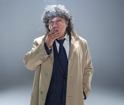 The senior man in cloak with cigar as detective or mafia boss. Studio shot on gray in retro stile